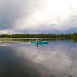 Person kayaking at Chena Lake Recreation Area. Forest and cloudy sky in background.
