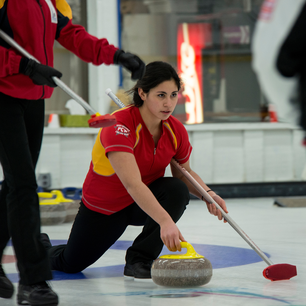 Curler in a team uniform sliding a stone on the ice during a curling match, with teammates nearby holding brooms.
