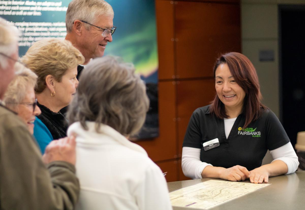 A smiling visitor center staff member at Explore Fairbanks assists a group of visitors, pointing to a map on the counter.