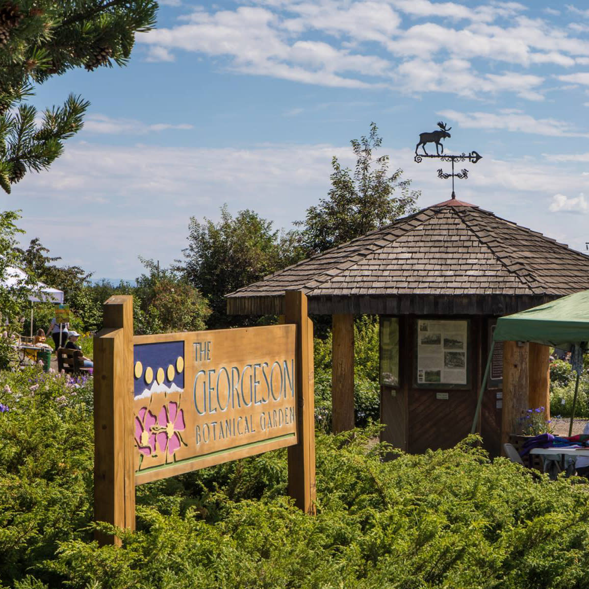 Wooden entrance sign for the Georgeson Botanical Garden surrounded by lush greenery and colorful flowers, with a rustic information kiosk in the background.