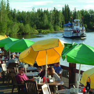 Patio along Chena River lined with yellow and green umbrellas and people lounging at tables.