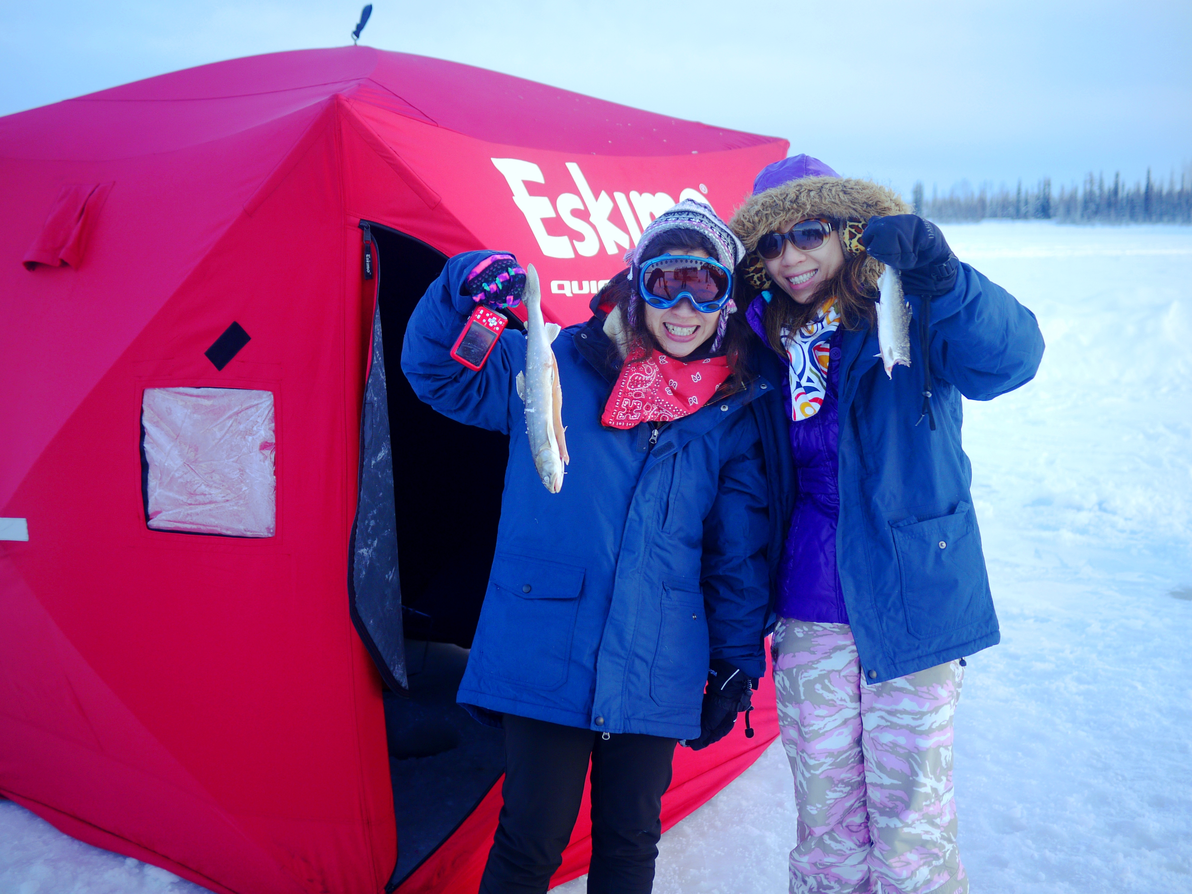 Two people holding up fish, standing in front of ice fishing tent
