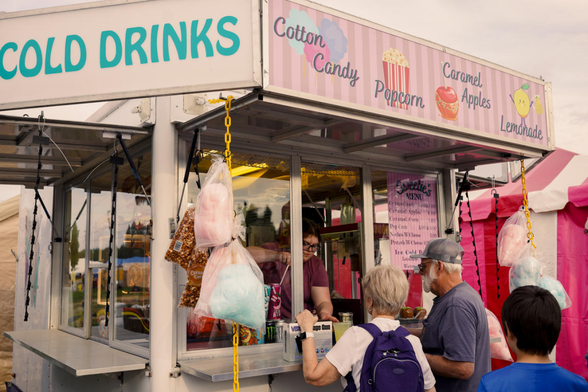 People ordering food from a fair food vendor. The vendor's sign advertises cotton candy, popcorn, caramel apples, and lemonade.