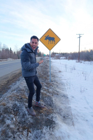 Reuben Mourad smiling next to a moose crossing road sign during an Explore Fairbanks Media Tour