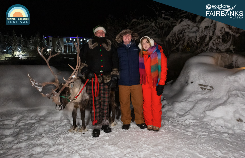 Three people in winter clothing smiling and standing next to a reindeer. Snow is on the ground and trees.